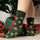 Dark green crew socks with a repeating pattern of green brussels Sprouts faces, posed in a holiday setting with wrapped presents and a Christmas tree.