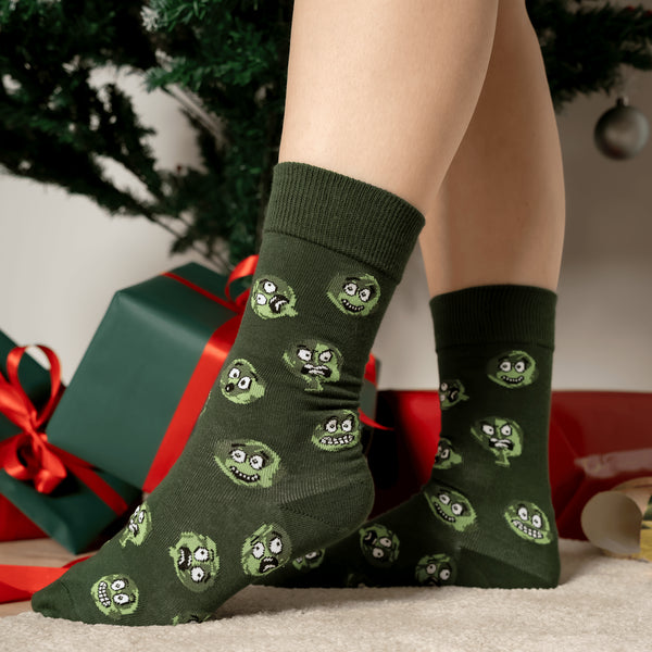 Dark green crew socks with a repeating pattern of green brussels Sprouts faces, posed in a holiday setting with wrapped presents and a Christmas tree.